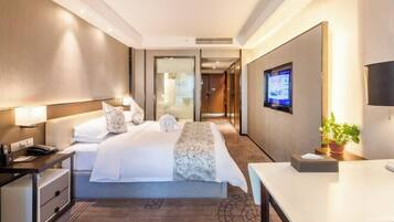 Quarto de hotel com cama, mesa de cabeceira, luminária, televisão e escrivaninha.