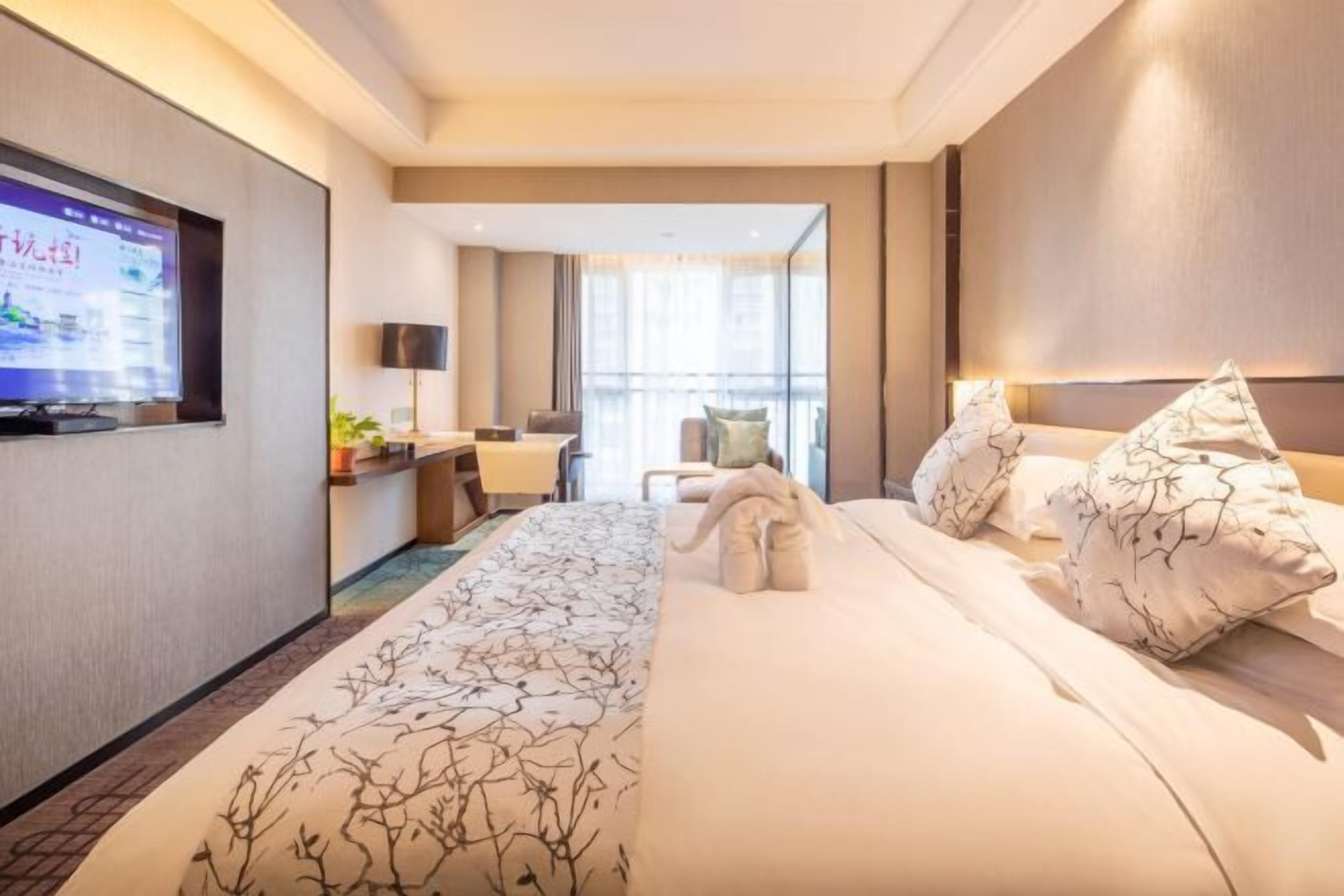 A hotel room with a large bed, a TV mounted on the wall, a desk with a chair, and a window with curtains.