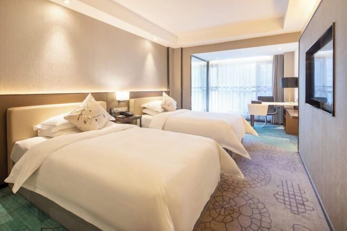 A hotel room with two beds, a desk, and a large window offering a view of the city.