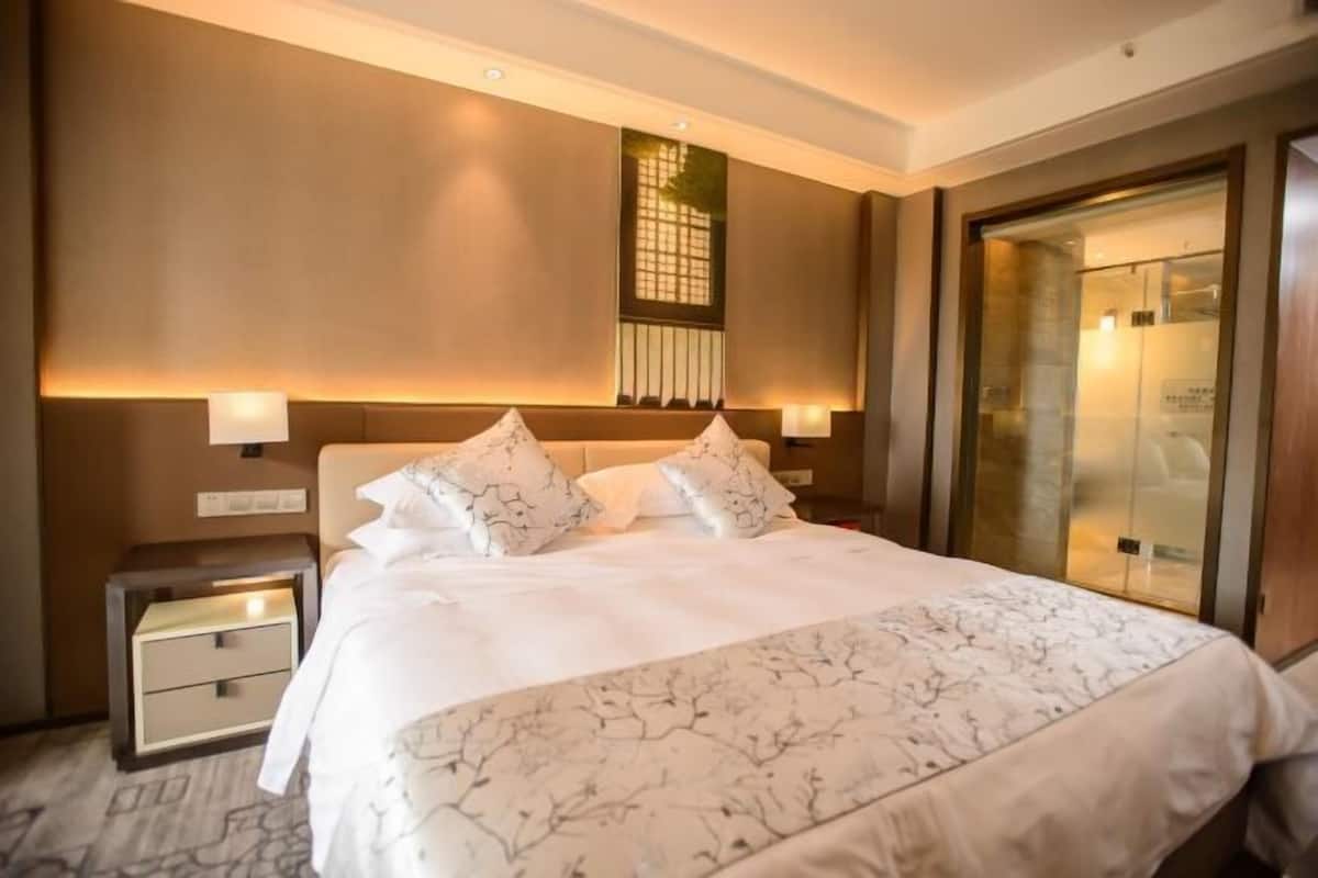 Ein Hotelzimmer mit einem großen Bett, Nachttischen, einem Beistelltisch, einem Fenster mit Jalousien und einer Tür mit Glasfenster.