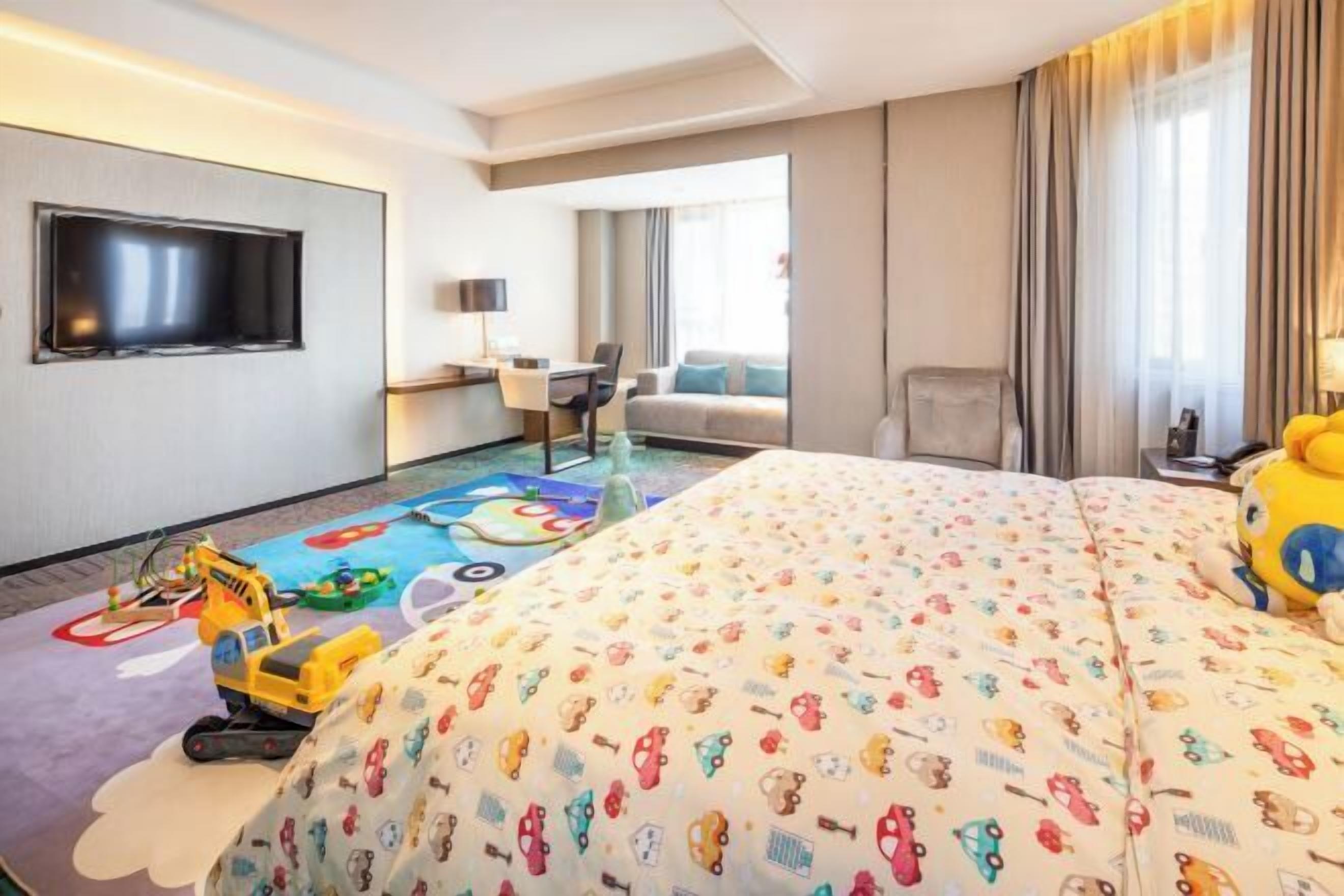 A hotel room with a large bed, a TV mounted on the wall, a desk with a chair, and a window with curtains.