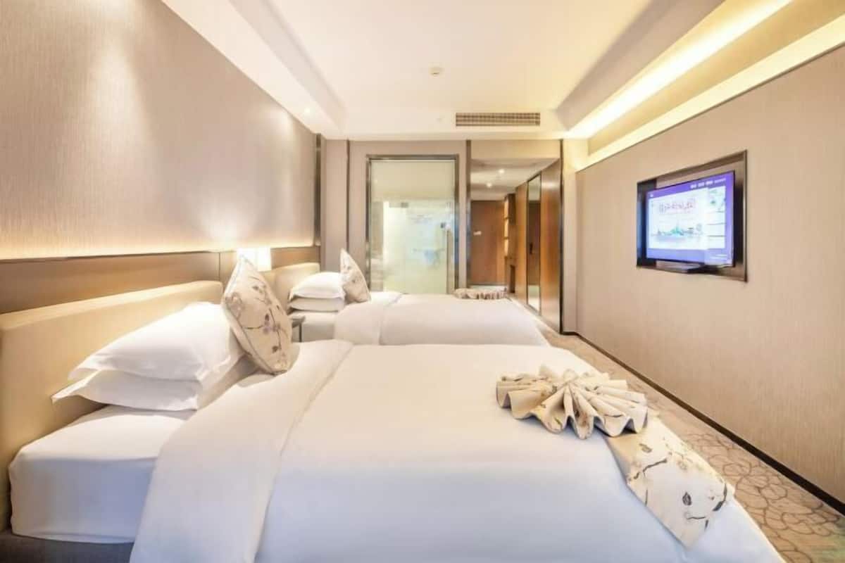 Ein Hotelzimmer mit zwei Betten, einem an der Wand befestigten Fernseher und einem durch eine Glastür einsehbaren Badezimmer.