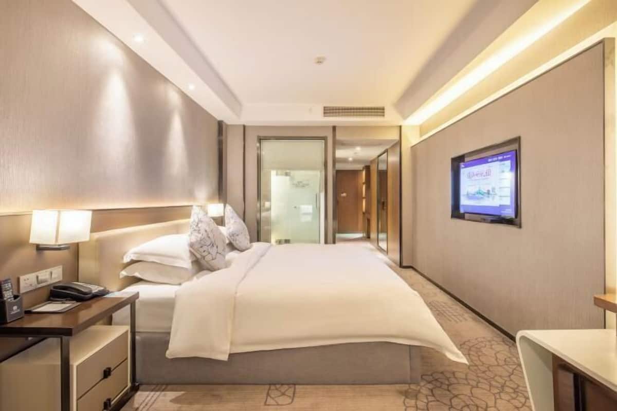 Ein modernes Hotelzimmer mit einem ordentlich bezogenen Bett, einem Nachttisch mit Lampe, einem flachbildfernseher an der Wand und einer Glastür zum Badezimmer.