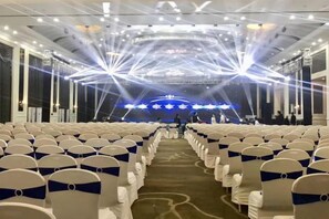 A conference room with rows of chairs, a stage with a blue backdrop, and a ceiling with light fixtures. - New Glory Holiday Inn (Jurong)
