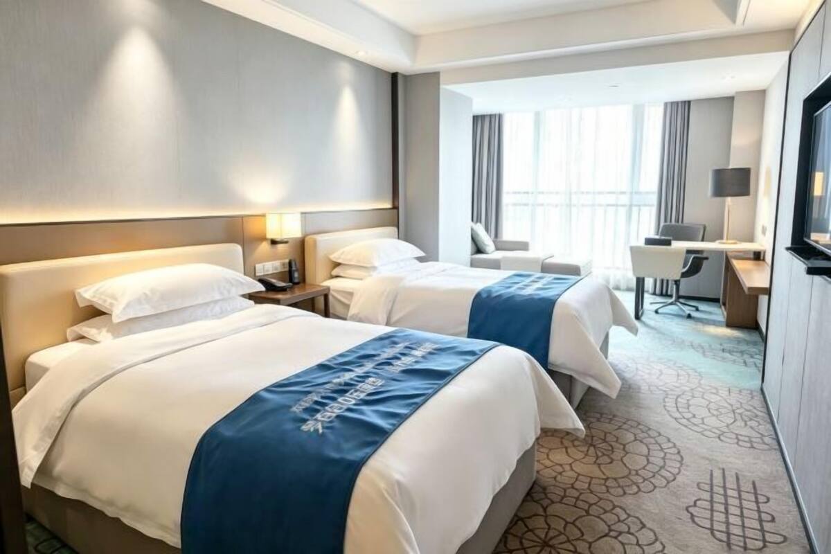 Ein Hotelzimmer mit zwei Betten, einem Schreibtisch, einem Sessel, einem Fernseher und einem großen Fenster.