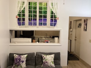 En stue med en sofa, to pyntepuder, et vindue med gardiner, og et køkkenområde med en vask og bordplade.