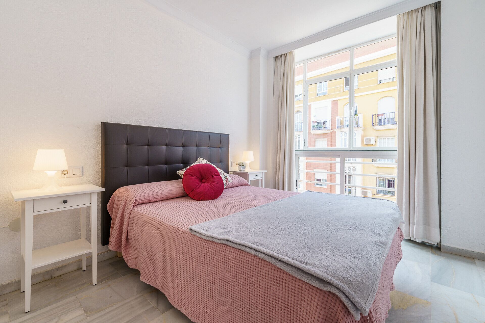 A bedroom with a large bed, two bedside tables, a window with a view of buildings, and a curtain.