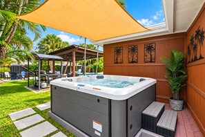 A hot tub in a backyard with a wooden fence, palm trees, and a covered patio area.