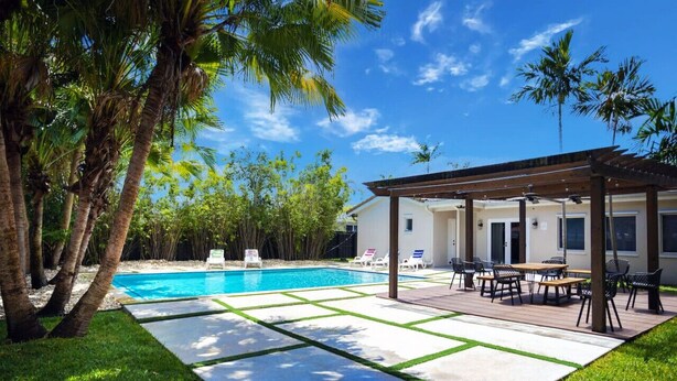 Ein Garten mit Schwimmbecken, Palmen, einer überdachten Terrasse mit Gartenmöbeln und einem Haus im Hintergrund.