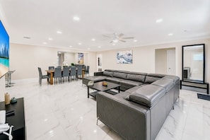 A modern living room with a sectional sofa, a dining area with a table and chairs, and a large flat-screen TV. - Deluxe 5BR/3BA Villa in Hollywood Lakes near Beach (Hollywood)