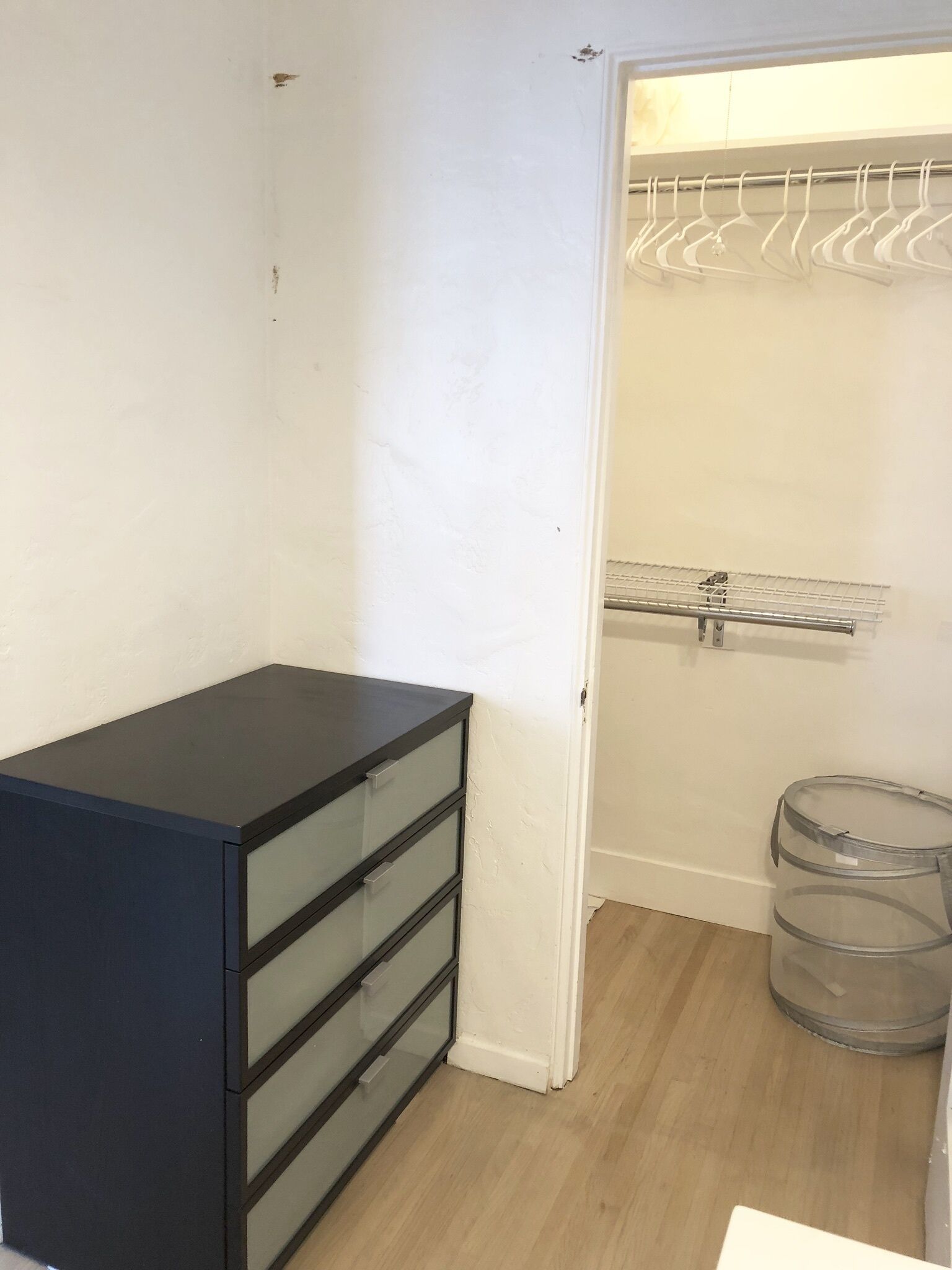 Une petite pièce avec une commode noire, un mur blanc, un plancher en bois, un placard avec un étendoir et une poubelle.