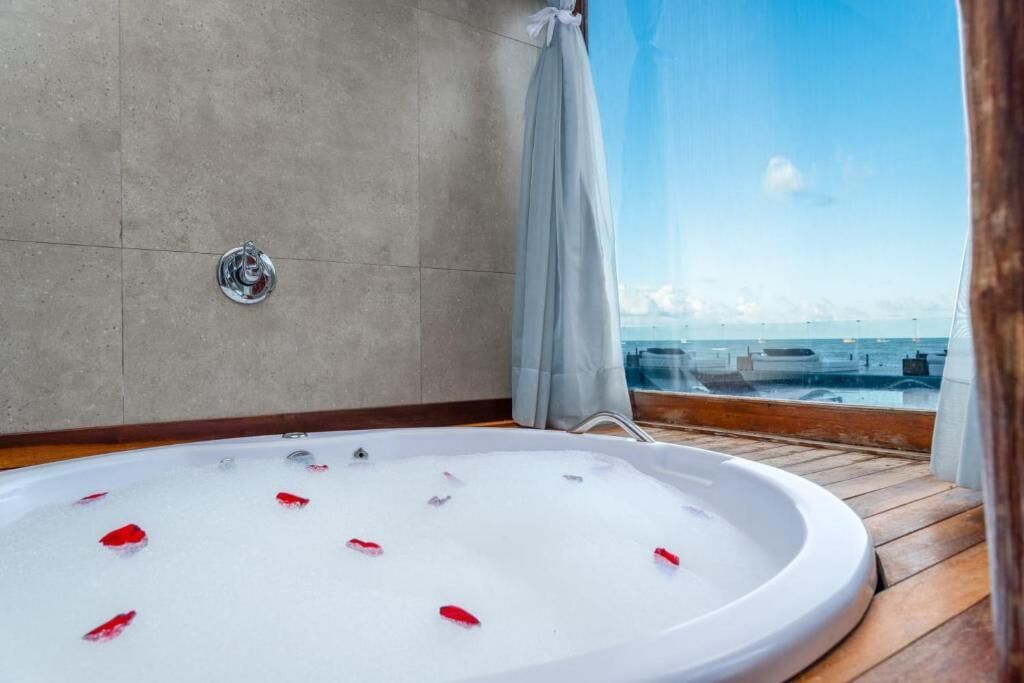Una bañera con pétalos de rosa flotando en el agua, una ventana con vista a barcos, y una cortina entreabierta.