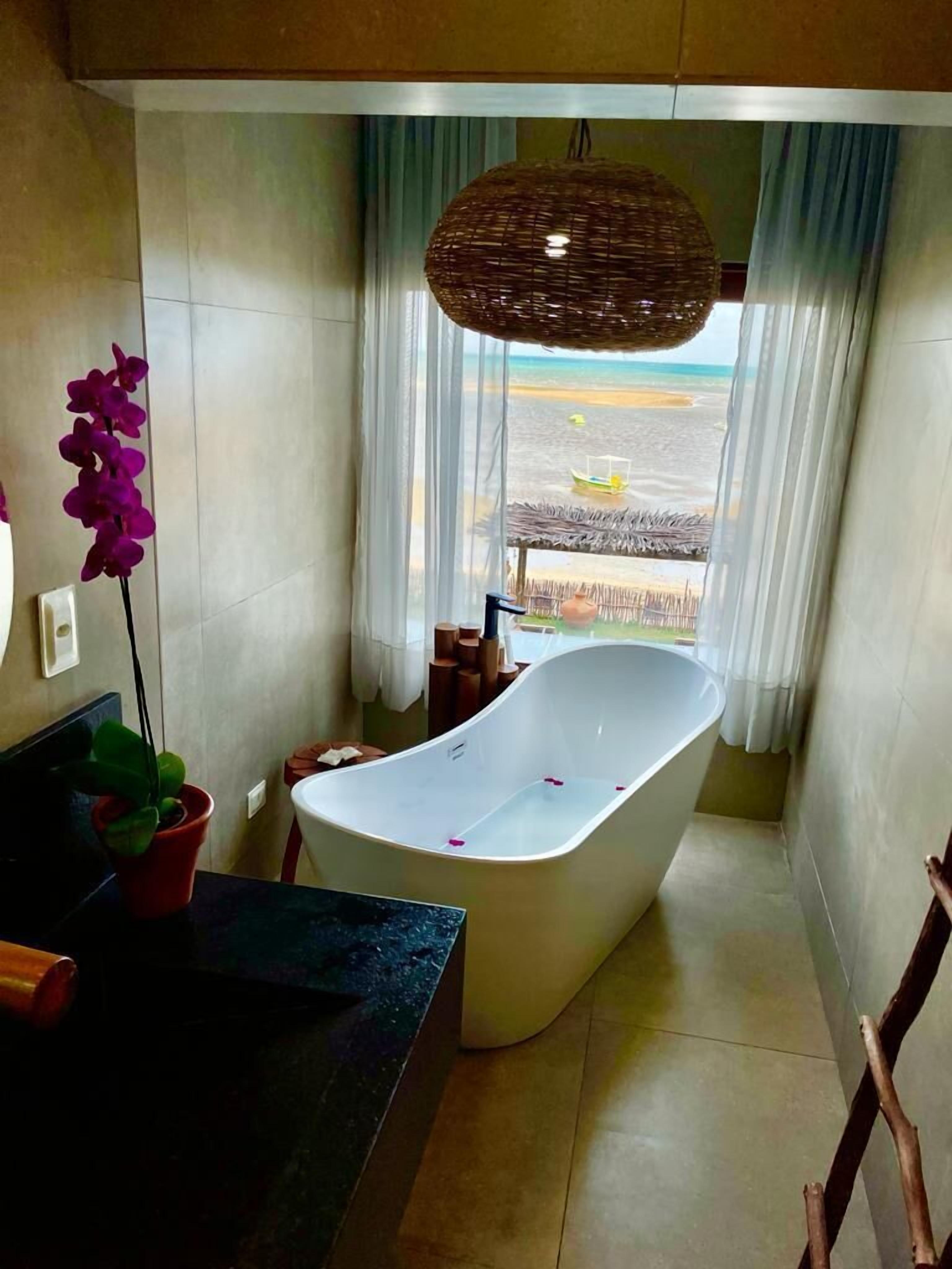 Un baño moderno con una bañera independiente, una planta en maceta y vistas a la playa a través de un ventanal amplio.