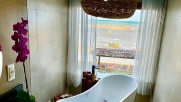 Un baño moderno con una bañera independiente, una planta en maceta y vistas a la playa a través de un ventanal amplio.