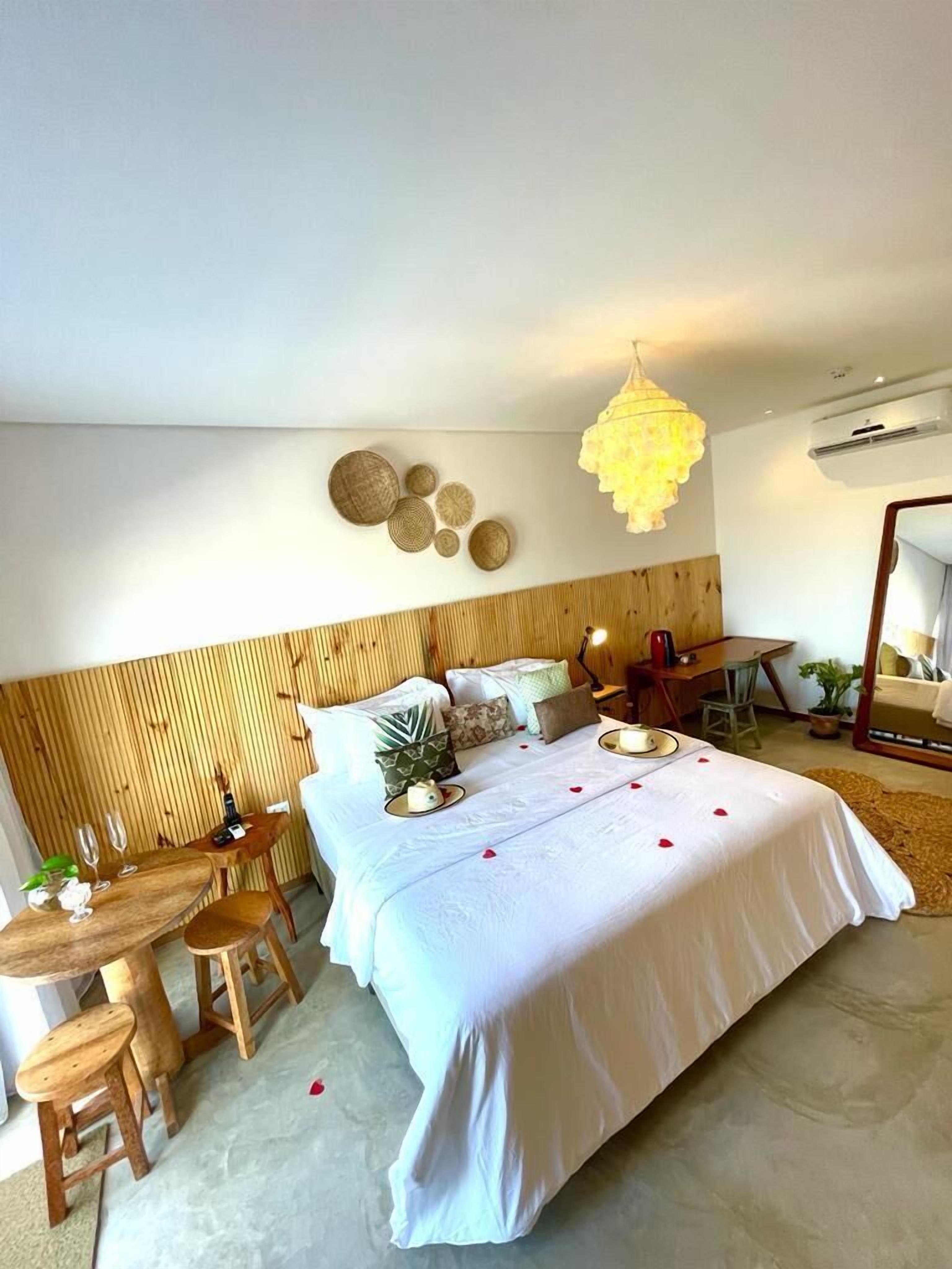 Habitación de hotel con cama, cabecero de madera, zona de comedor con mesa y sillas, y lámpara colgante.