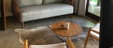 A living room with a grey sofa, a round wooden coffee table, a woven wall hanging, and a view through a glass door.