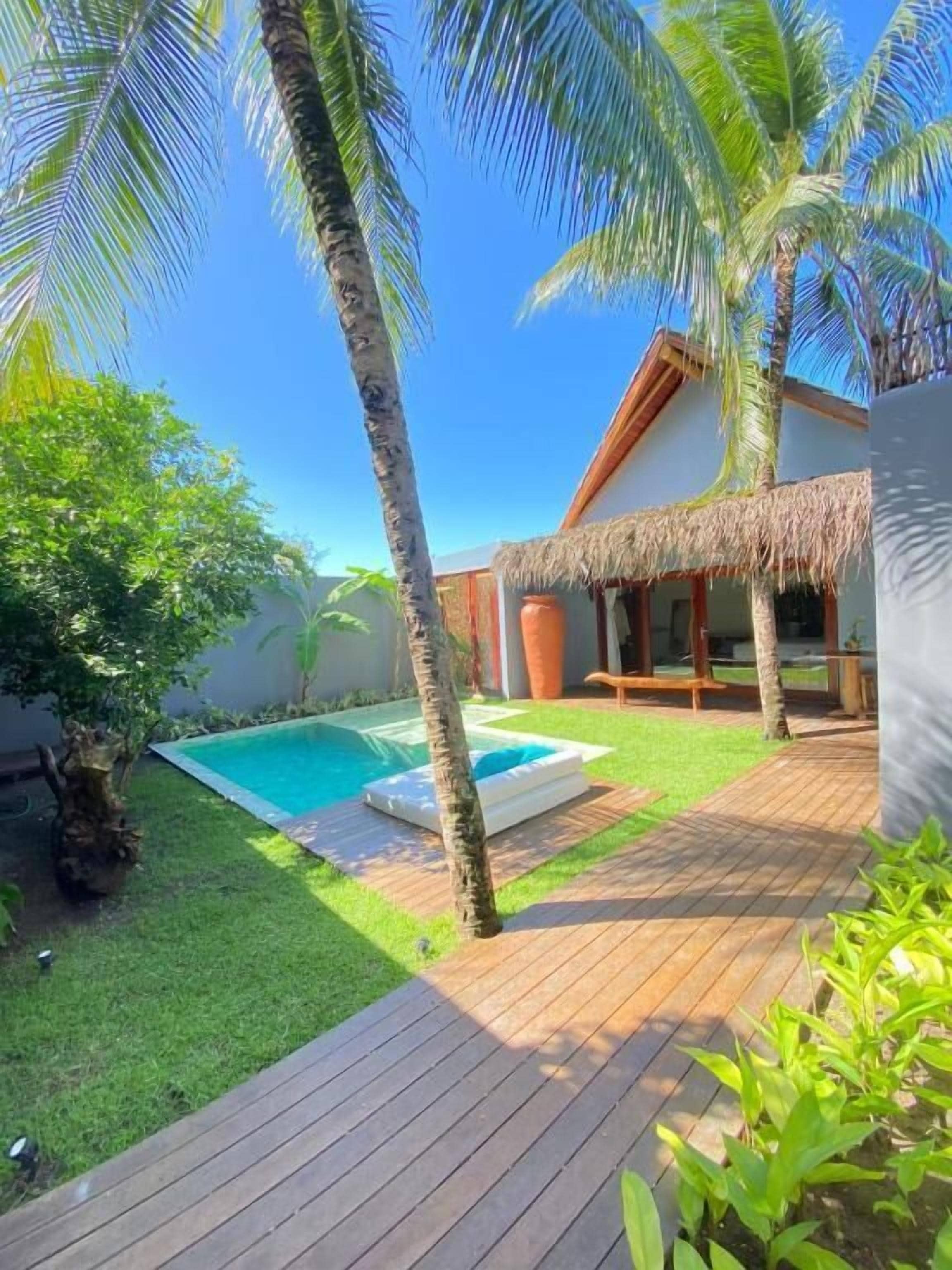 A pool area with a wooden deck, a thatched-roof structure, and palm trees.
