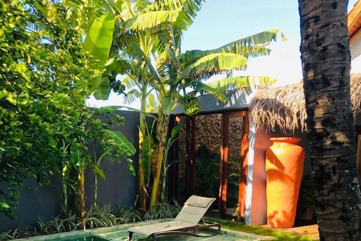 Ein rechteckiger Swimmingpool mit klarem, blauem Wasser, umgeben von einer Holzterrasse. Im Hintergrund befinden sich zwei Liegestühle und eine strohgedeckte Konstruktion.