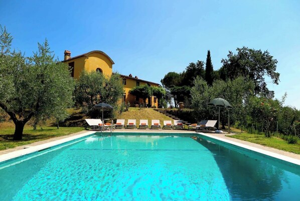 A swimming pool with lounge chairs and umbrellas, surrounded by trees and a building in the background.