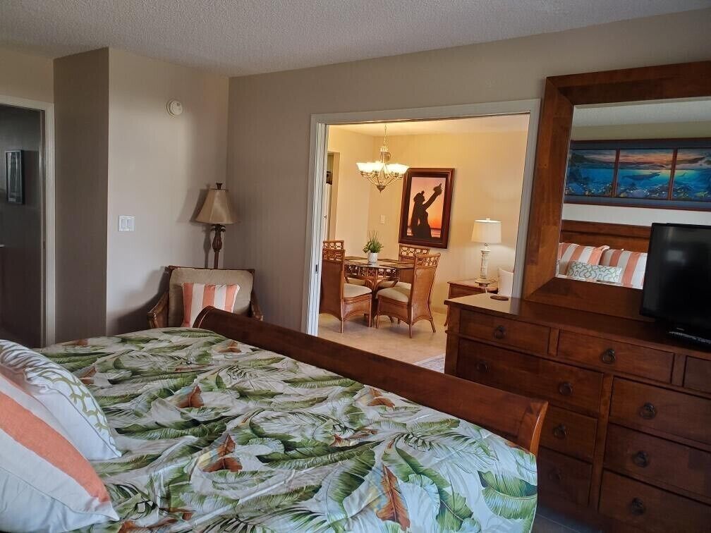 A hotel room with a bed, a dresser, a television, and a dining area visible through an open door.