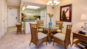 A dining area with a round glass table surrounded by wicker chairs, a chandelier, and a framed picture on the wall. - Maui Banyan ground floor 2 full baths,steps to Kamaole 2,great walking location. (Kihei)