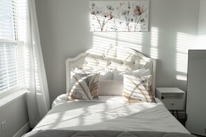 Quarto com cama, mesa de cabeceira, janela com persianas e uma obra de arte na parede.
