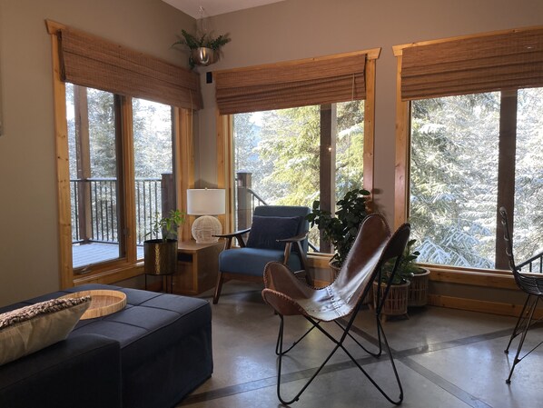 A cozy living room with a blue sofa, a brown leather chair, and a small table with a lamp. Large windows with wooden frames offer views of snow-covered trees.