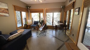 Un vaste salon doté d’un ventilateur de plafond, un coin repas avec une table et des chaises, et de grandes fenêtres offrant une vue sur les arbres.