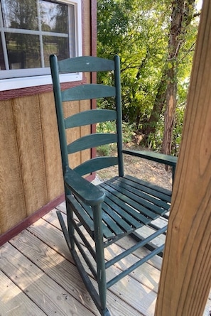 Ein grüner Metall-Schaukelstuhl auf einer Holzdielen-Terrasse mit Fenster und Grünpflanzen im Hintergrund.