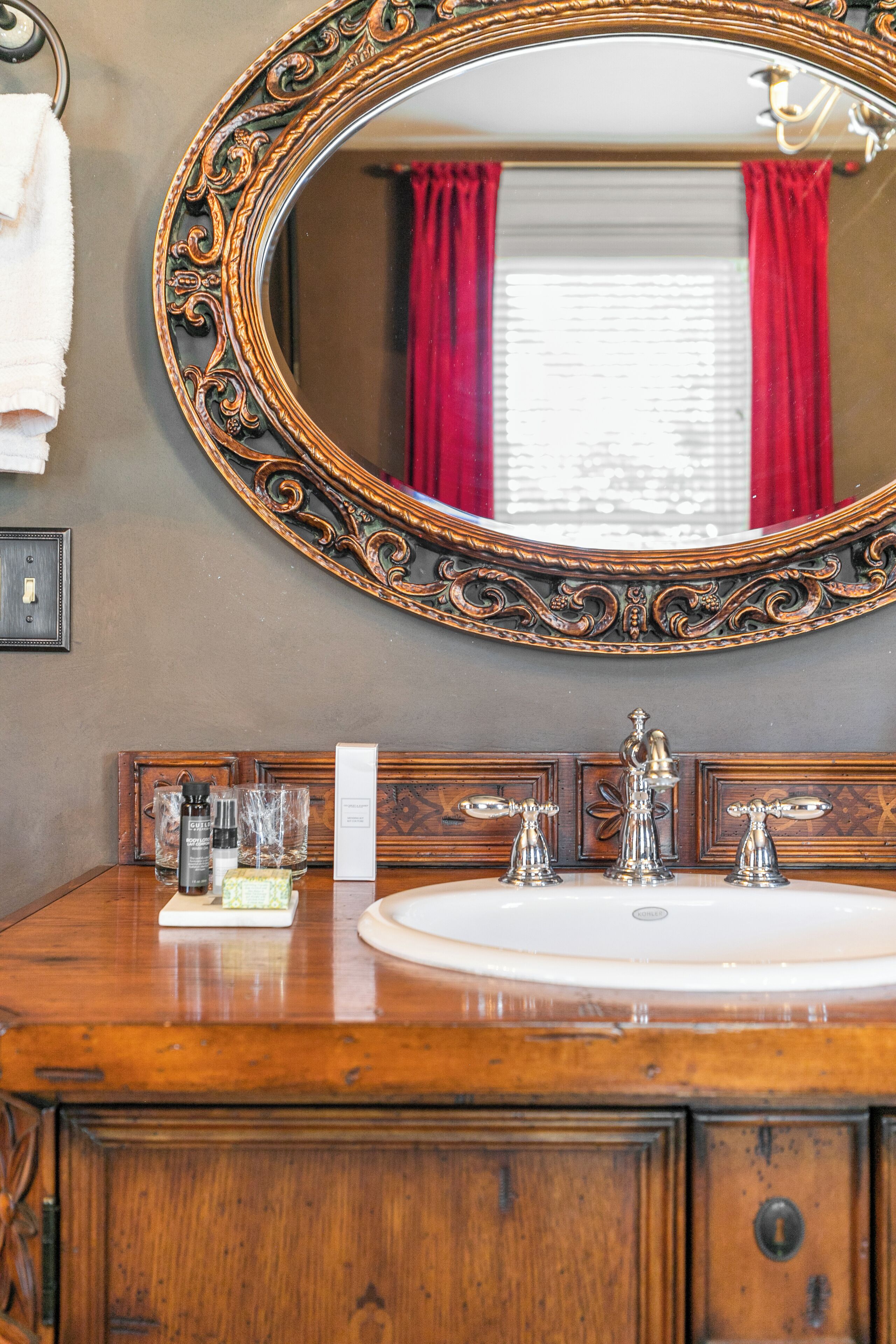 A wooden vanity with a large ornate mirror, a white sink, and toiletries.