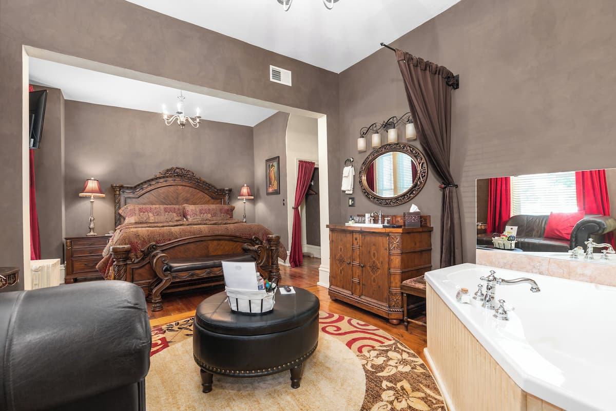 A bedroom with a large bed, a wooden dresser, a sitting area with a chair, a desk, and a large window with curtains.