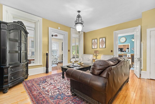 A spacious living room with a brown leather sofa, a patterned rug, a dark wooden cabinet, and a ceiling light fixture.