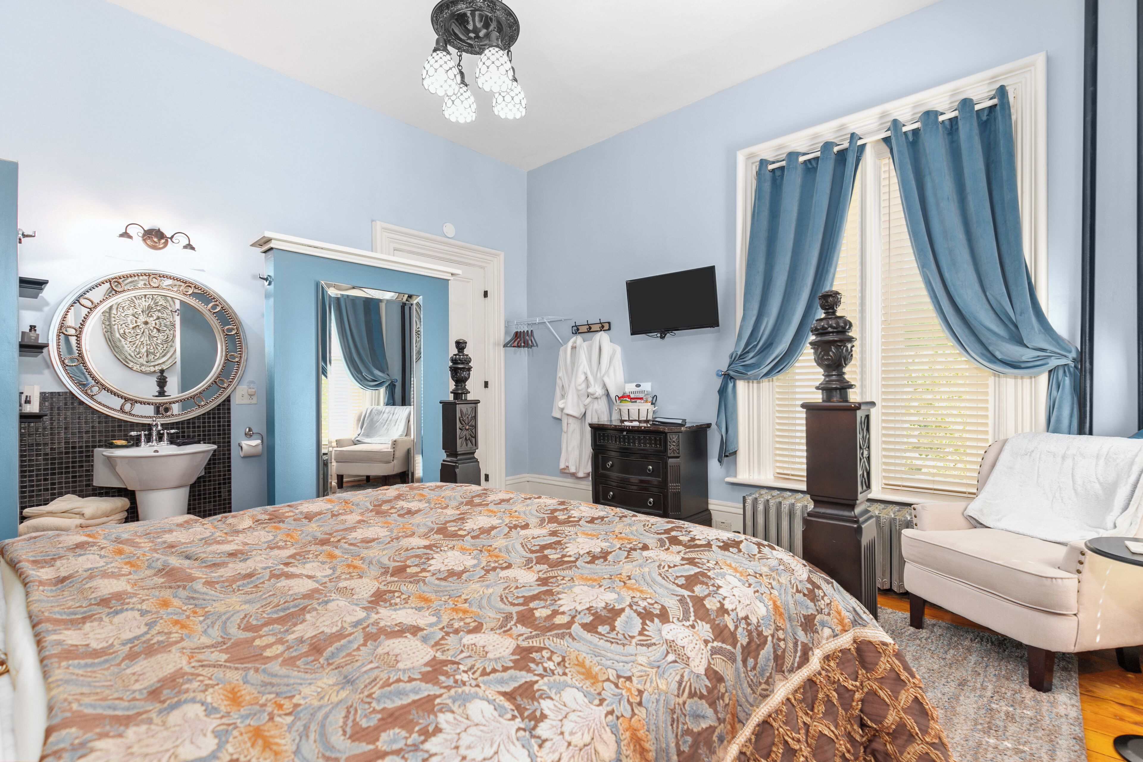 Un dormitorio con una cama grande, un aparador, una tocador con espejo, un televisor y una ventana con cortinas azules.