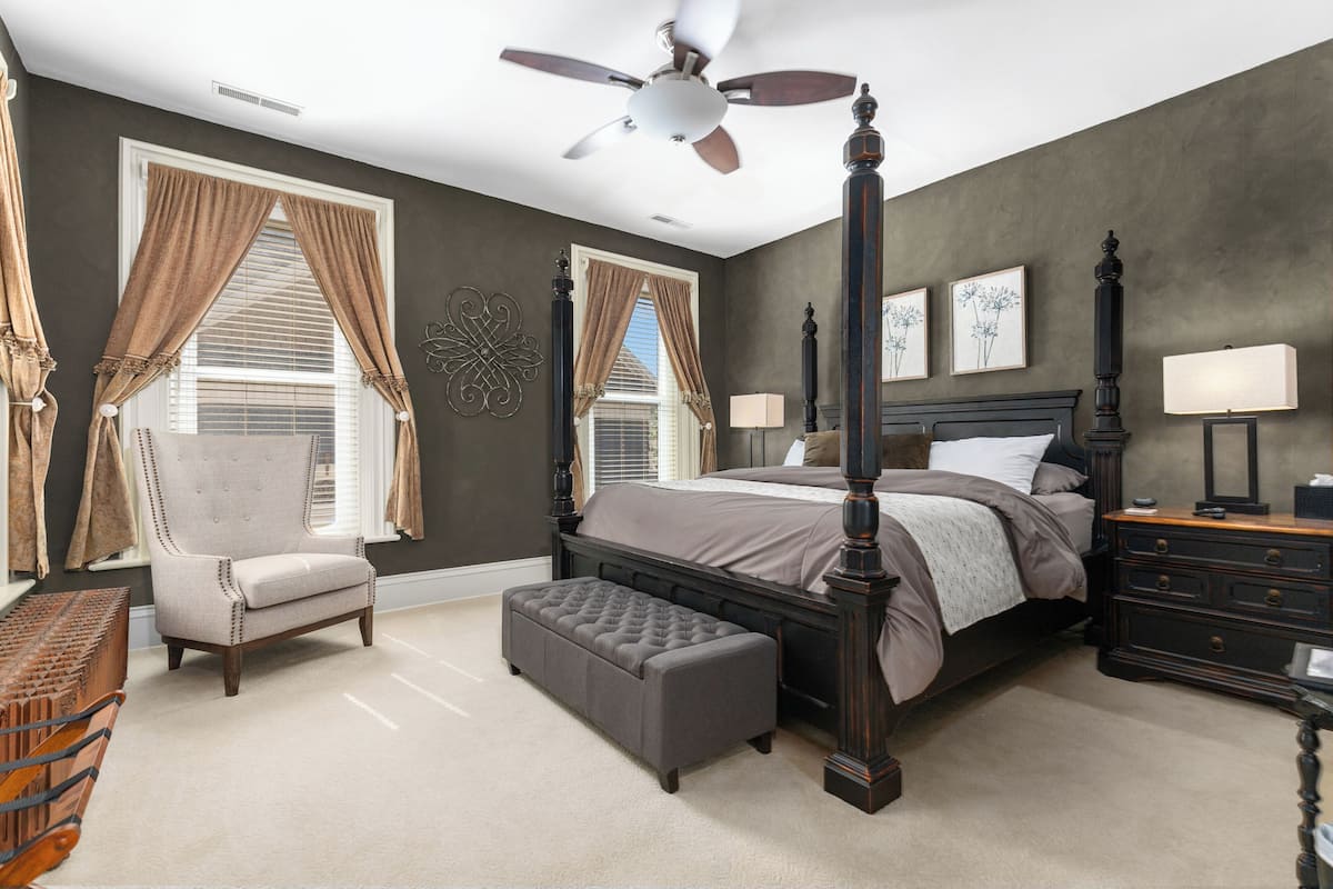 A bedroom with a four-poster bed, a chair, an ottoman, a nightstand, and a ceiling fan.