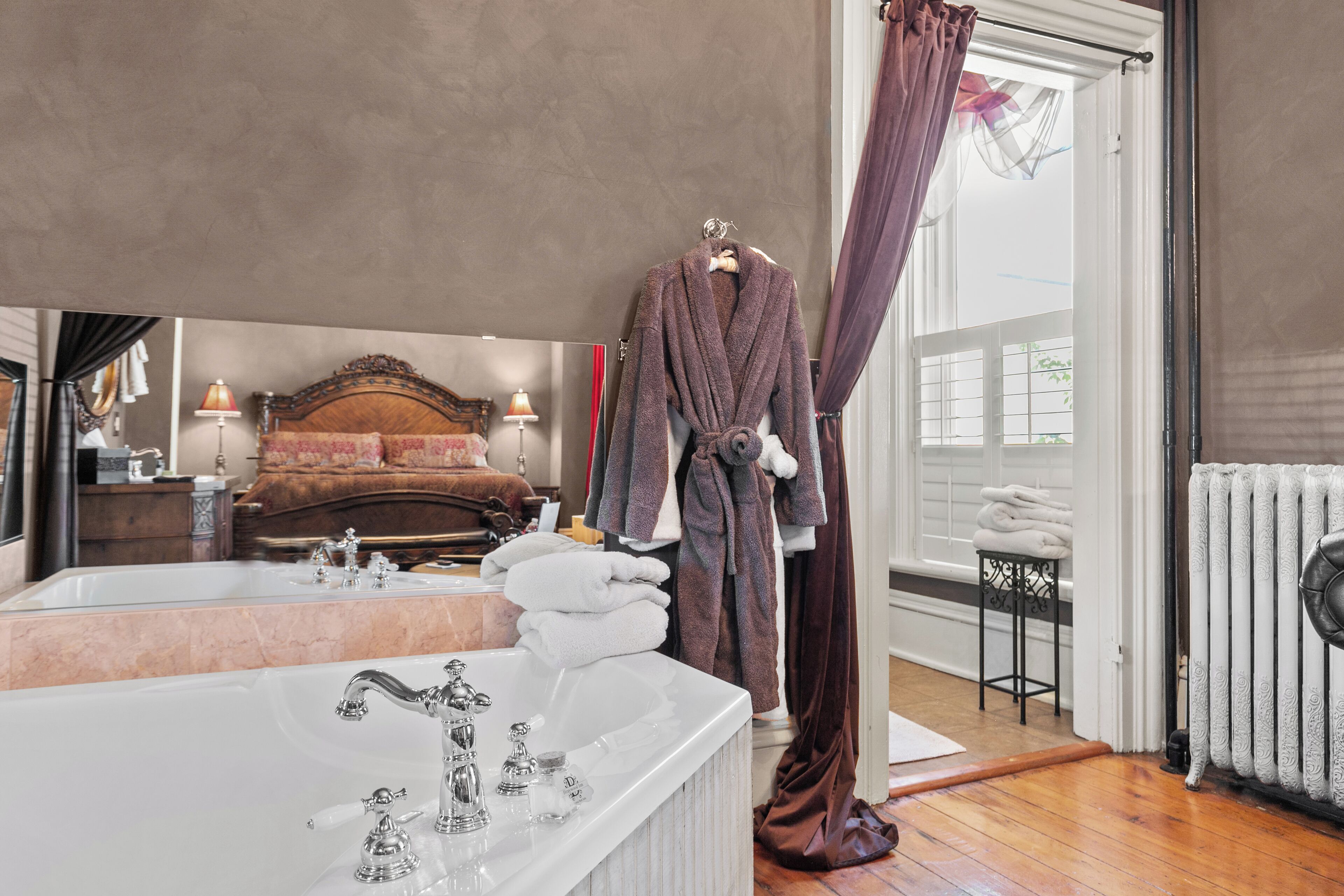 A hotel room with a large mirror, a white bathtub, a wooden bed, and a window with curtains.