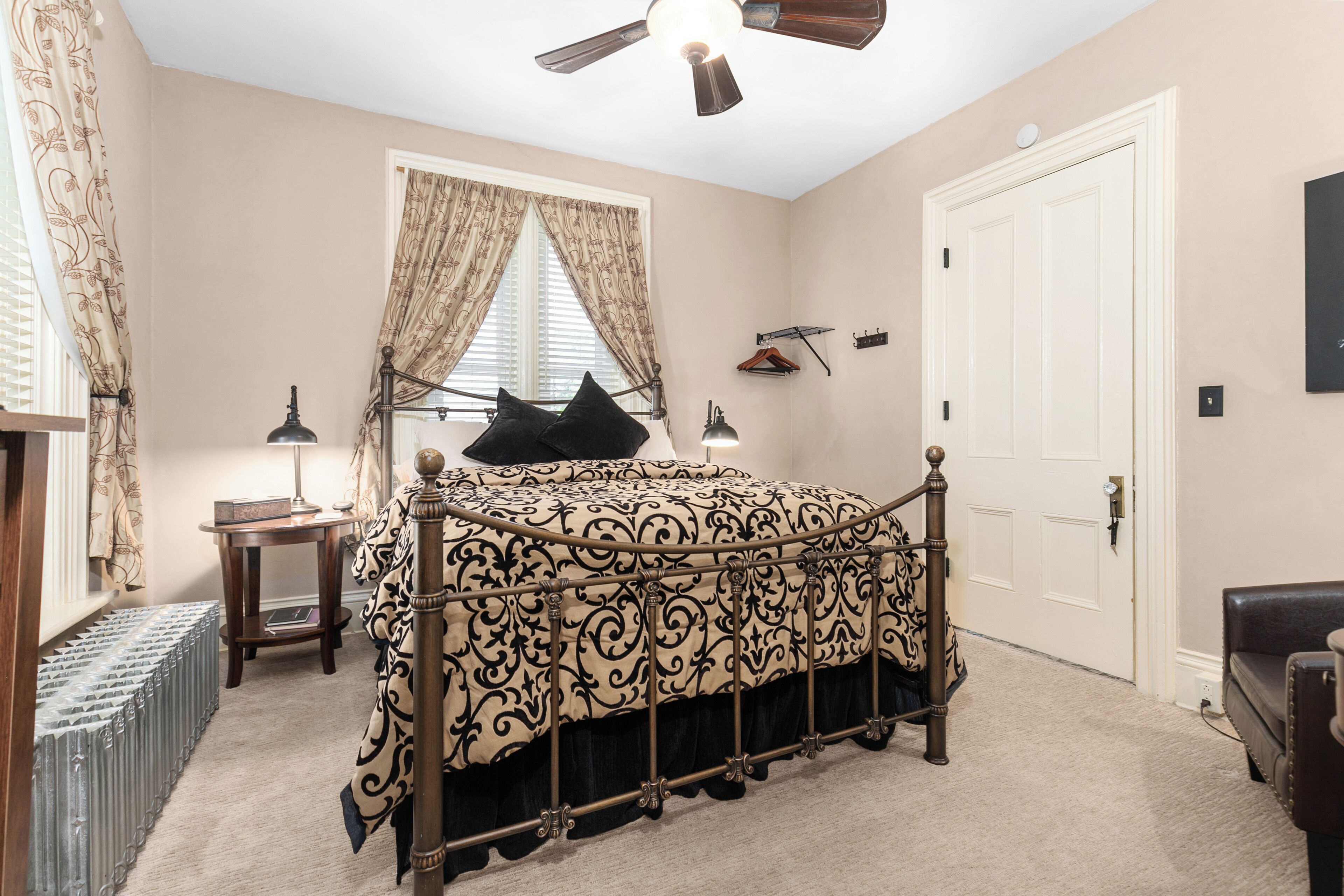 A bedroom with a bed, a ceiling fan, a window with curtains, a door, and a coat rack.