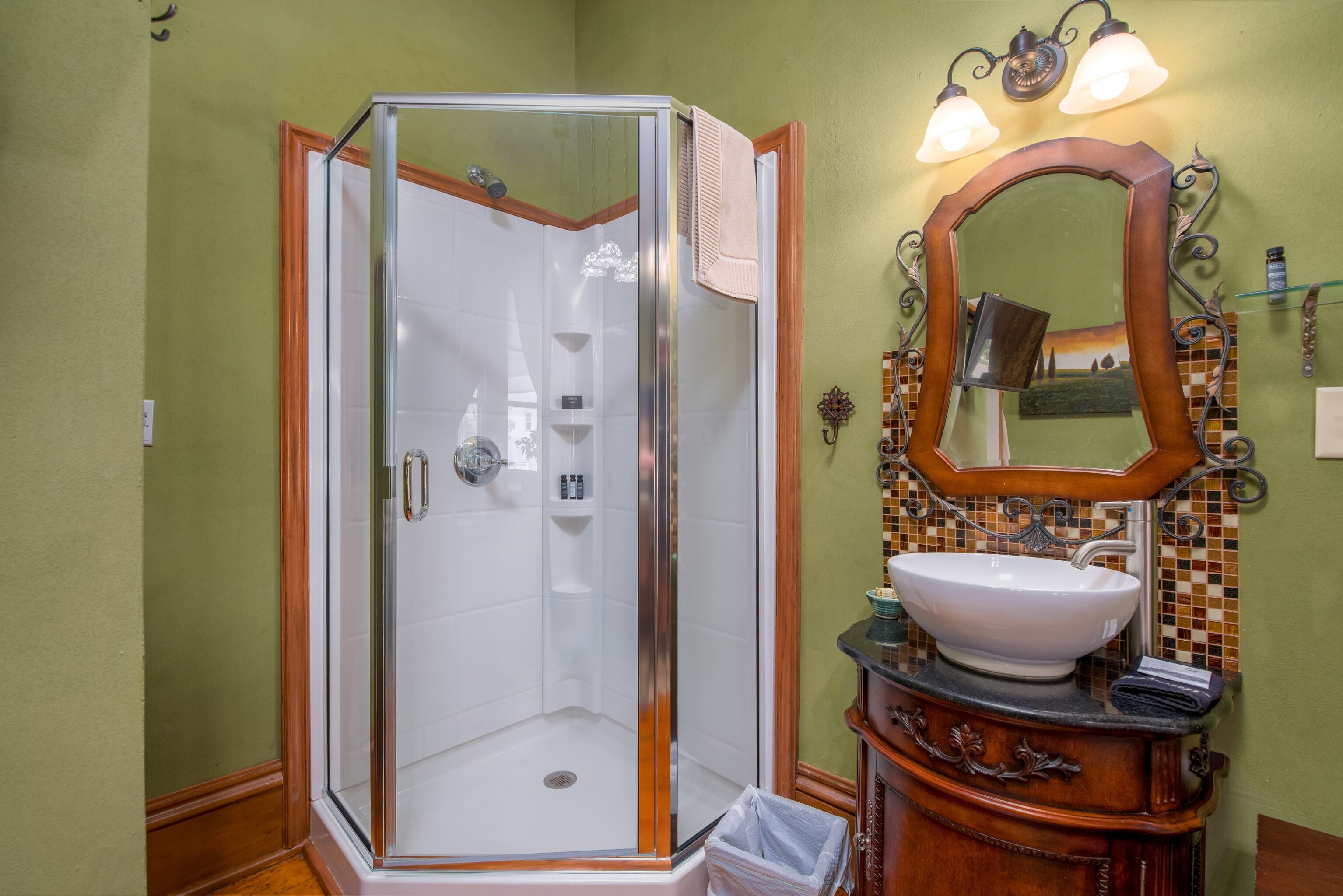 Un baño con ducha acristalada, tocador de madera con lavamanos y espejo con luces decorativas.