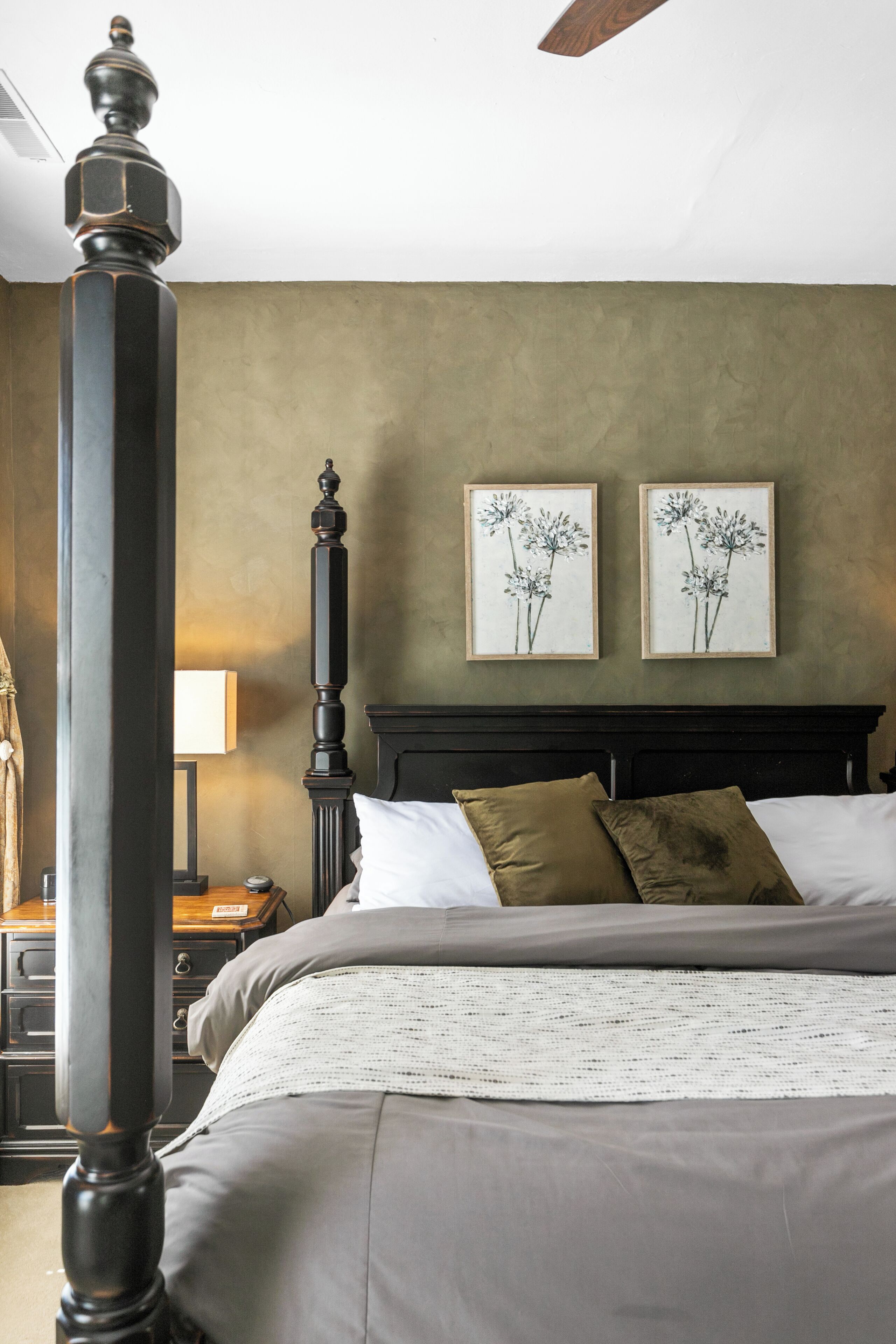 A bedroom with a four-poster bed, a nightstand, a lamp, and two framed floral paintings on the wall.