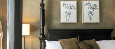 A bedroom with a four-poster bed, a nightstand, a lamp, and two framed floral paintings on the wall.