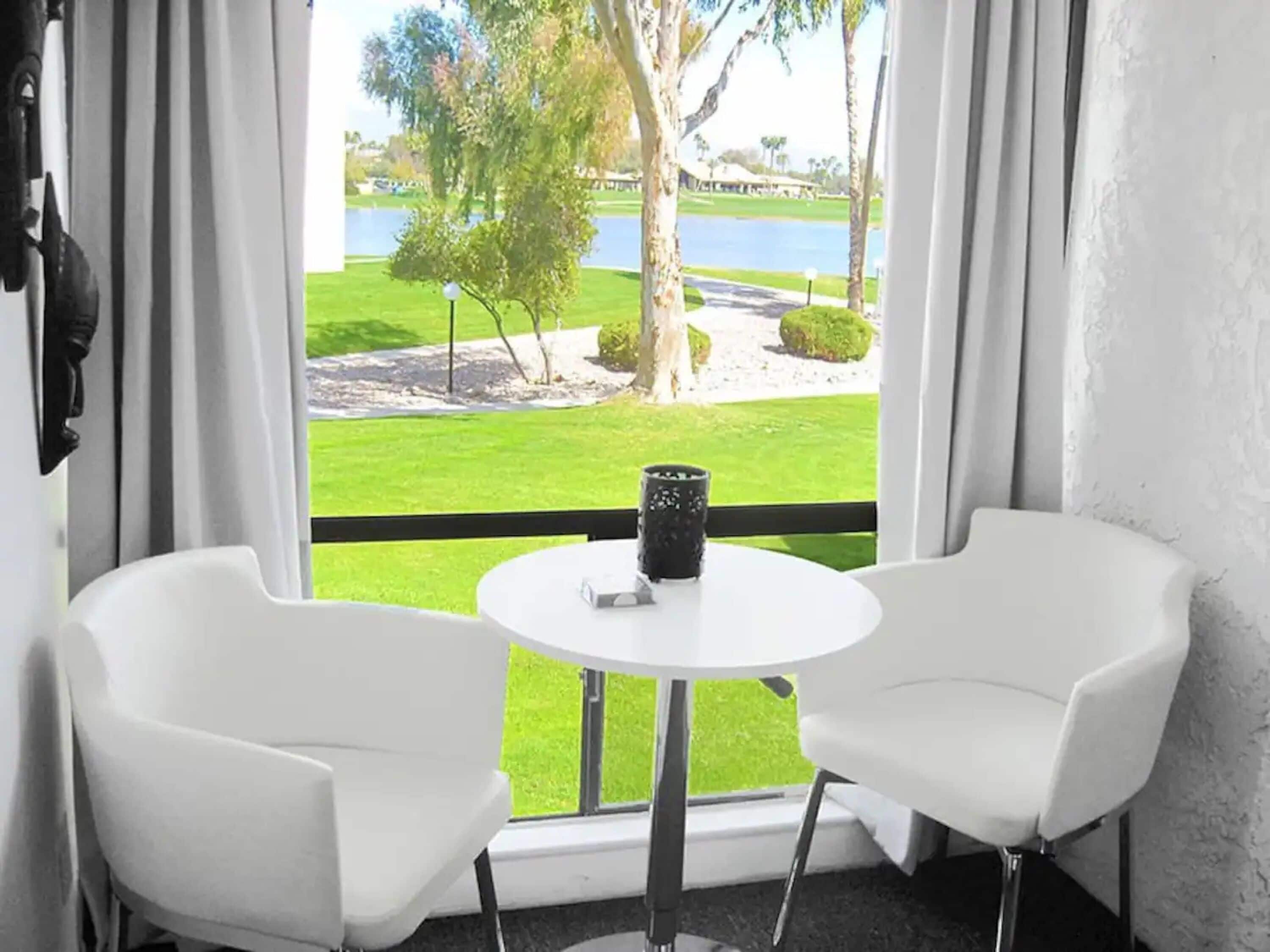 A room with two white chairs and a small table with a black object, overlooking a well-maintained lawn and a body of water.
