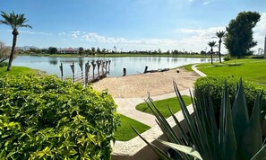 A serene lakeside park with a sandy beach, a walking path, and lush greenery.