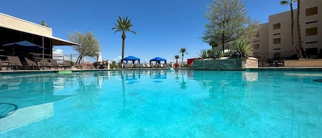 Una piscina azul cristalina con sillas de descanso y sombrillas, rodeada de palmeras y un edificio con varias ventanas.