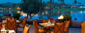 Un espacio para comer al aire libre con mesas y sillas, una piscina y vista a un campo de golf al anochecer.
