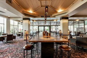 A spacious indoor dining area with a central wooden table surrounded by chairs, large windows, and a high ceiling with exposed beams.