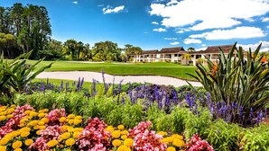 Un parcours de golf avec un bunker de sable, entouré de fleurs aux couleurs vives et de plantes tropicales, et un bâtiment au style architectural traditionnel en arrière-plan.