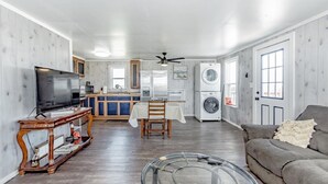 A spacious living room with a grey sofa, a wooden coffee table, and a television. A dining area with a table and chairs is visible in the background.
