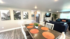 Un coin repas avec une table en bois, des chaises blanches et des sets de table orange. On aperçoit une cuisine en arrière-plan, avec un réfrigérateur et un téléviseur fixé au mur.