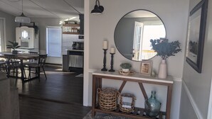 A modern living room with a dark wood floor, a dining area with a wooden table and chairs, a kitchen with white cabinets and a built-in oven, and a hallway with a round mirror and decorative items on a console table. - Lake view retreat steps to Badin Lake! Pool, Beach, Restaurant/Bar & Boat Launch (New London)