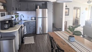 A modern kitchen with grey cabinets, a stainless steel refrigerator, a coffee machine, and a dining area with a wooden table and chairs. - Lake view retreat steps to Badin Lake! Pool, Beach, Restaurant/Bar & Boat Launch (New London)