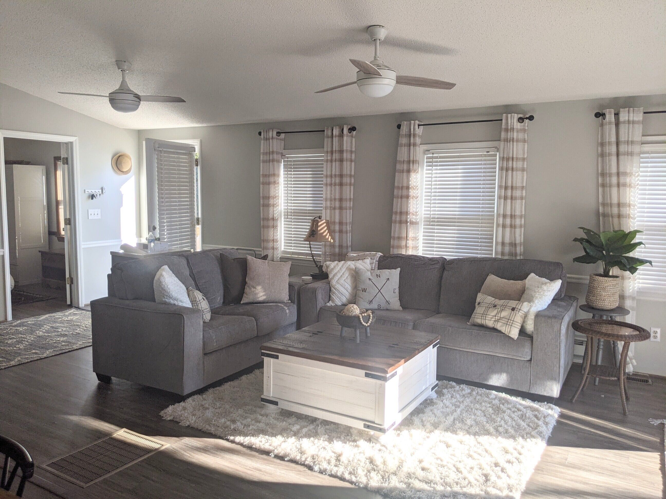 A living room with a grey sectional sofa, a white coffee table, and a potted plant. There are two ceiling fans, a window with blinds, and a door leading to another room.